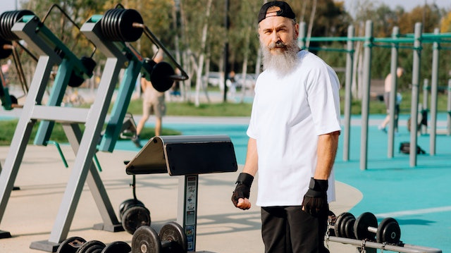Man with a long beard in a black baseball cap and fingerless gloves standing next to some gym equipment